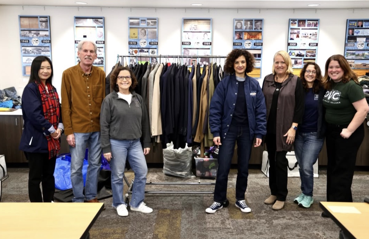Seven smiling volunteers stand beside a rack of donated coats and suits for the Community Closet clothing drive
