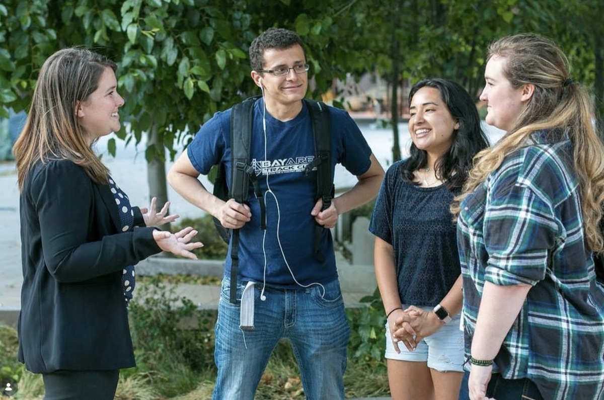 Rebecca Barrett talks with three smiling college students outdoors among green trees on campus