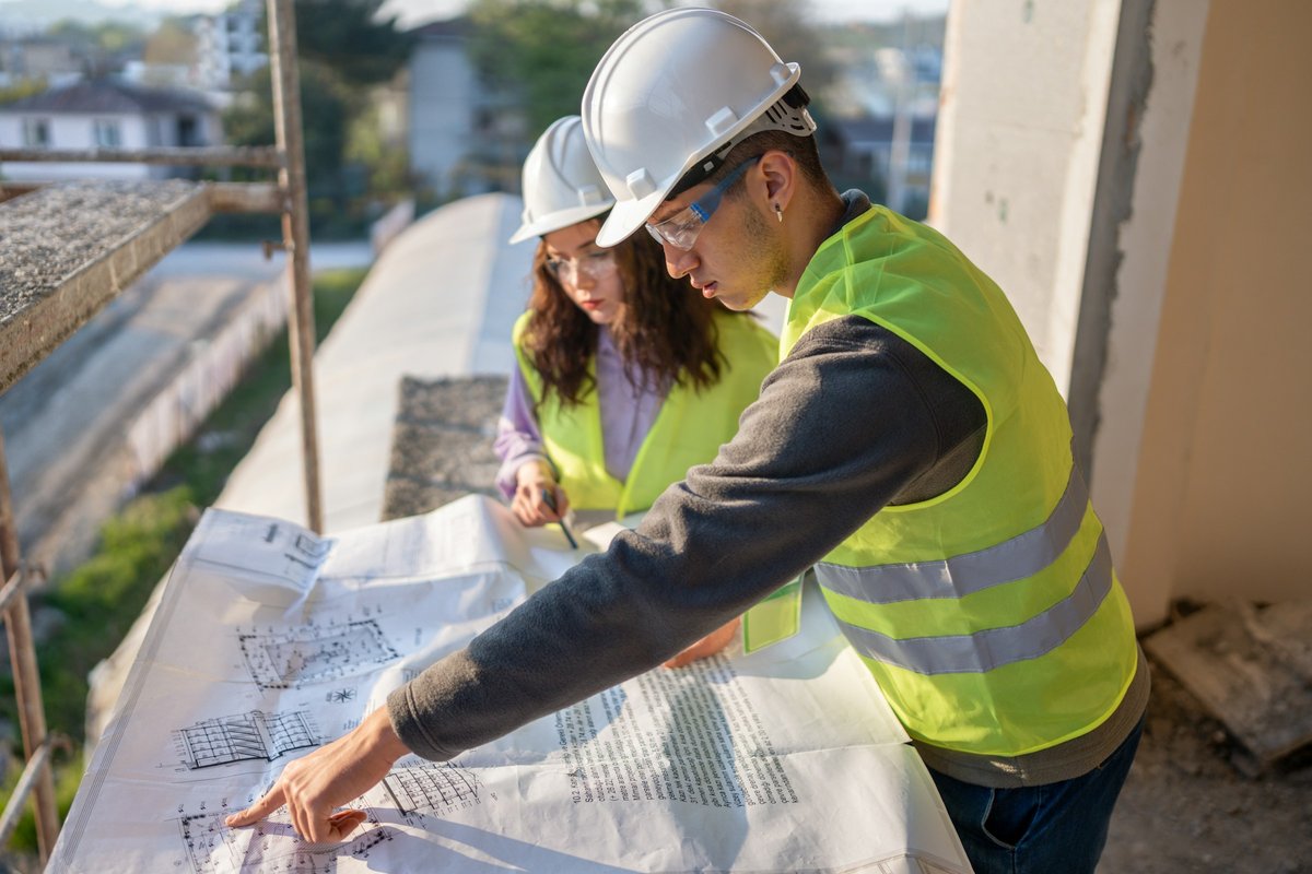 Two young construction workers in hard hats and safety vests review blueprints on a job site