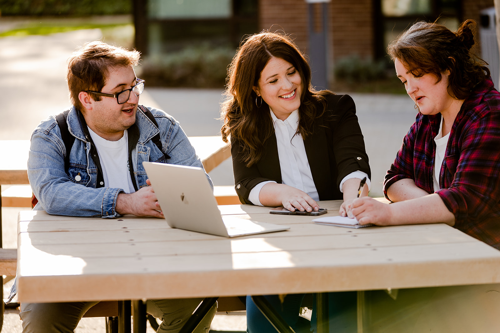 Rebecca Barrett sits at an outdoor campus picnic table with two students, one with a laptop and one taking notes
