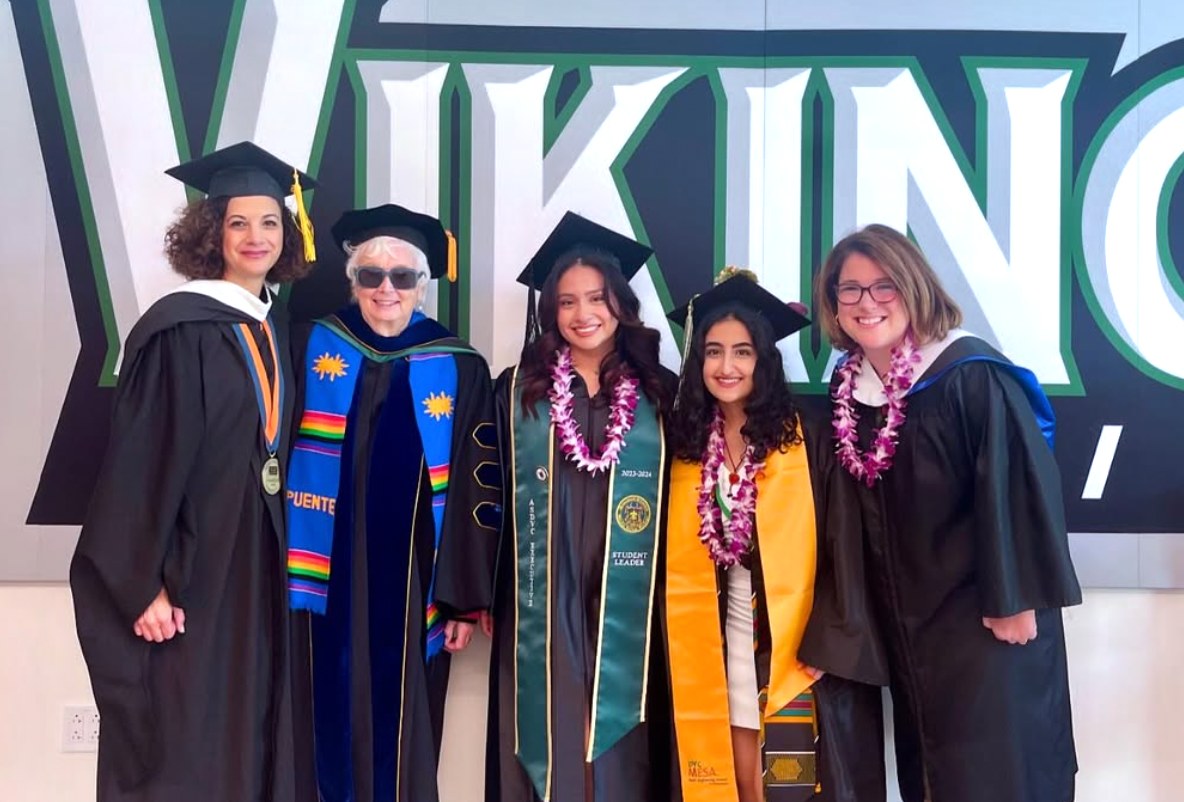 Rebecca Barrett in academic regalia with four graduates wearing stoles and leis in front of a DVC Vikings backdrop