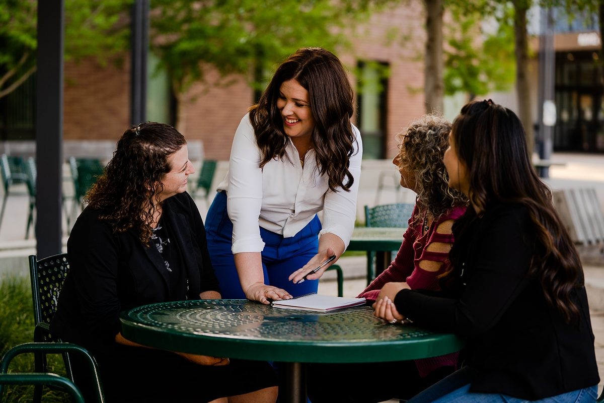 Rebecca Barrett leans over a patio table in conversation with three smiling women on campus